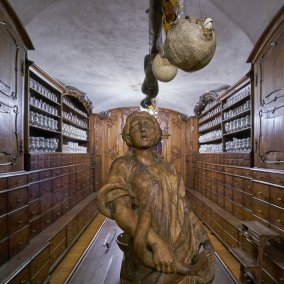 Holzgeschnitzte Figur auf einem Apothekentisch, links und rechts das Mobiliar der Innsbrucker Hofapotheke. An der Decke hängt ein ausgestopfter Kugelfisch