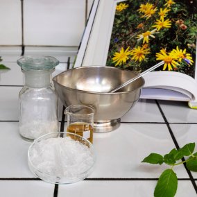 Utensilien um ein eigenes Gel herzustellen, Petrischale, Metallgefäss, Glas mit Arnikaöl, dahinter eine aufgeschlagene Buchseite mit der Arnikablume