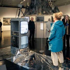 Besucher:innen der Ausstellung stehen vor zwei übereinander gestapelten Fernsehern, auf denen ein Video läuft.