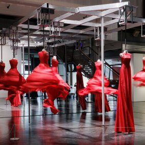 Besucher:innen betrachten rote Kleider, die von der Decke hängen und sich drehen.