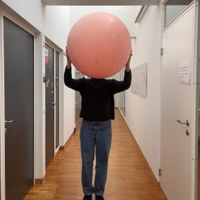 Person hält in einem Gang rosa Gymnastikball vor ihr Gesicht.