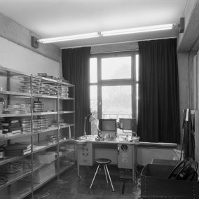 Schwaez-weiß-Fotografie: Raumansicht eines Büros mit einem Regal an der linken Wand und einem Schreibtisch vor einem Fenster.