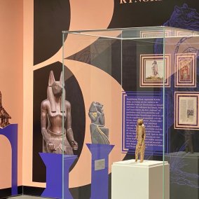 Farbfotografie: Ansicht eines Ausstellungsraumes mit farbiger Wantapete. Im Zentrum eine Vitrine, in der auf einem weißen Sockel eine braune Figur steht.