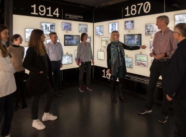 Der Ausstellungsraum zum 19. Jahrhundert zeigt eine Wand mit digitalen, historischen Bildern. Davor steht eine Menge von Personen, die sich unterhalten.