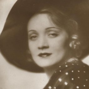 Die Fotografie zeigt ein Detail einer Fotografie der Schaupspielerin Marlene Dietrich. Die Fotografie ist schwarz-weiß. Dietrich blickt direkt in Richtung Betracher*in.
