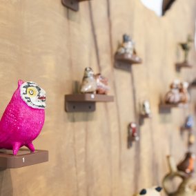Auf einem Baumstamm an einer Wand stehen viele kleine Eulen auf kleinen Podesten. Im Vordergrund sitzt eine pinke Eule.