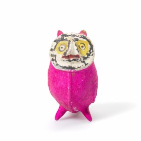 Eine kleine pinke Figur mit bemaltem Gesicht steht vor hellem Hintergrund. Sie wirkt wie eine handgemachte Kunst- oder Spielzeugfigur.