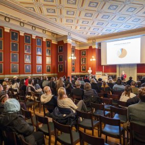 Blick in die Aula des Naturhistorischen Museums Basel, bestzte Stuhlreihen, Blick auf die Leinwand.