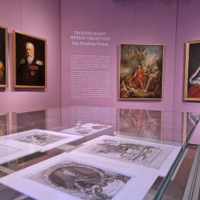 Blick in die Ausstellung mit Gemälden an der Wand und Grafiken in den Vitrinen