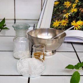Utensilien um ein eigenes Gel herzustellen, Petrischale, Metallgefäss, Glas mit Arnikaöl, dahinter eine aufgeschlagene Buchseite mit der Arnikablume