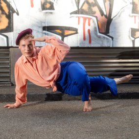 Person mit lachsfarbener Bluse, blauer Hose und rotem Hut macht am Boden eine Pose aus dem Voguing