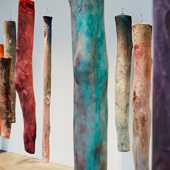 Das Bilde zeigt einen Blick in die Installation des Werkes Mothers leg's der Künstlerin Kaari Upson. An Drahtseil hängen baumstammartige Elemente von der Decke. Bei näherem betrachten, erinnern sie an ein Ausschnitt von Beinen mit Knien. Verfremdet wird die Darstellung durch das bunte Erscheinungsbild der Elemente.