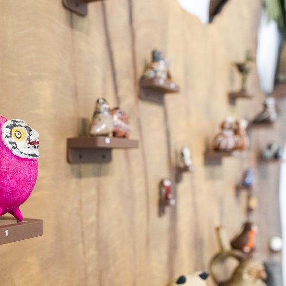 An einem braunen Baumstamm stehen auf kleinen Podesten ganz viele Eulen. Die Eule im Vordergrund ist pink.