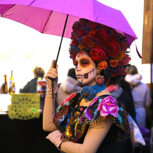 Catrina, üppig geschminkte Frau mit Rosen in den Haaren und sehr farbigem Kleid in rosa, orange, elb, blau, grün