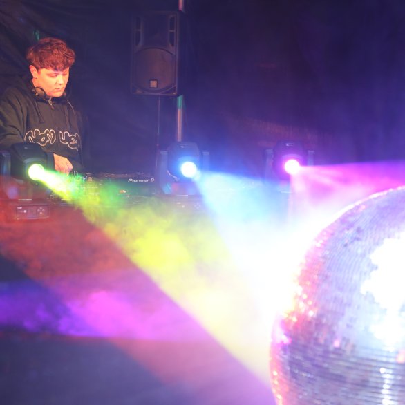 Djane an den Turntables und vor ihr strahlen die Lichter in Regenbogenfarben auf eine Discokugel und alles reflektiert farbenfroh