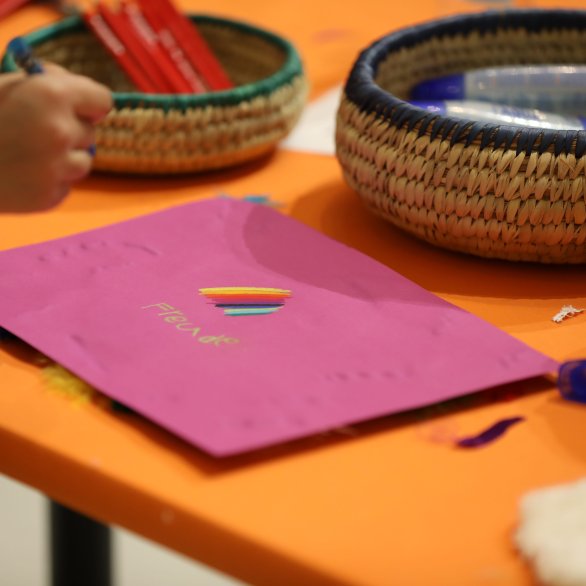 Auf einem orangen Tisch liegt ein pinkes Blatt, auf dem eine Kinderhand zeichnet.