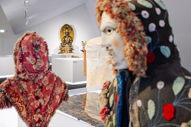 Zwei Masken "Rölli mit Kapuze"  aus dem Kanton  Schwyz von ca. 1900 – im Vordergrund zwei Masken, eine von hinten, mit rotem Stoff und die andere von der Seite mit aufgenähtem Blumenmuster auf schwarzem Stoff und einer plüschigen Kapuze. Im Hintergrund der goldene, grosse Buddha