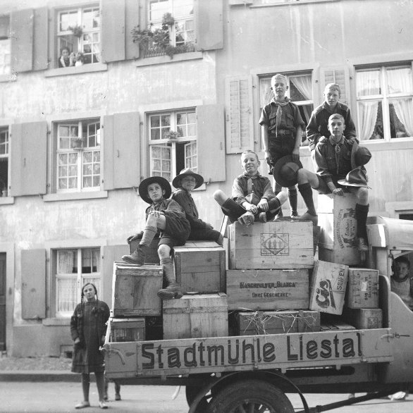 Auf der Schwarzweissfotografie von 1927 ist ein alter Lastwagen mit der Aufschrift "Stadtmühle Liestal" zu sehen, auf dessen Ladefläche sich Obstkisten stapeln. Auf und um den Wagen stehen und sitzen acht Kinder und Jugendliche in Pfadiuniform, die meisten grinsen stolz in die Kamera. Im Hintergrund sieht man Häuser, eine Frau schaut aus dem Fenster.