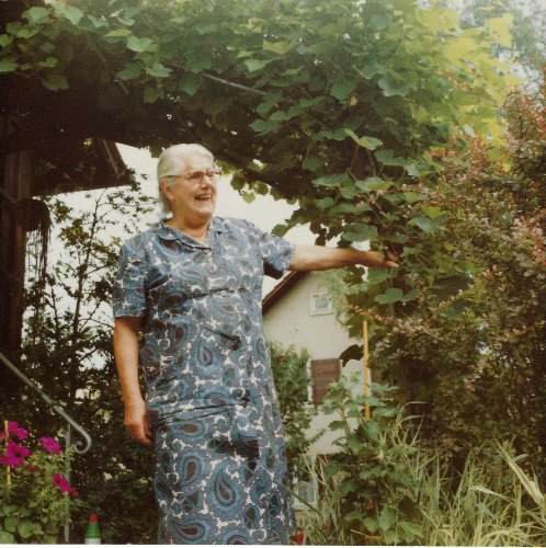 Zu sehen ist eine ältere, lachende Frau mit weissem Haar und Brille, umgeben von grünen Pflanzen.