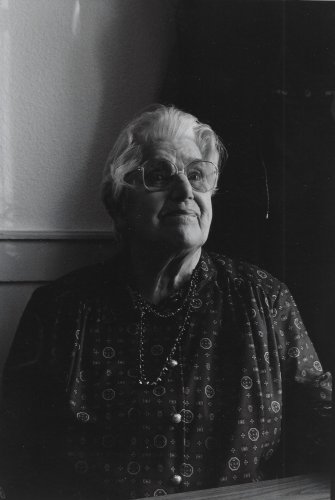 Die Schwarzweissfotografie zeigt eine ältere, weisshaarige Frau mit Brille, die ernst blickt.