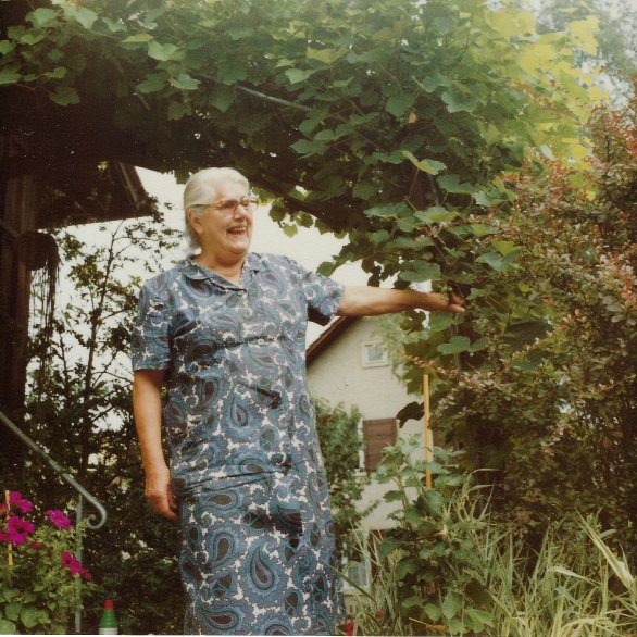 Zu sehen ist eine ältere, lachende Frau mit weissem Haar und Brille umgeben von grünen Pflanzen.