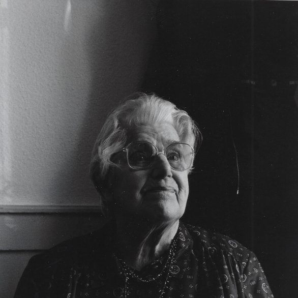 Die Schwarzweissfotografie zeigt eine ältere, weisshaarige Frau mit Brille, die ernst aussieht.
