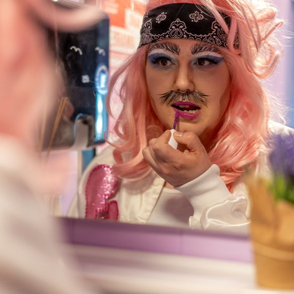 Eine geschminkte Person mit pinker Perücke sieht in einen Spiegel und trägt Lippenstift auf.