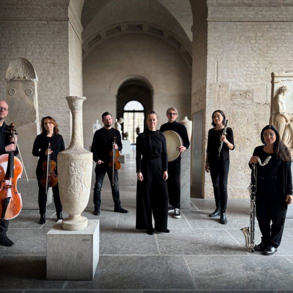 Sieben Musiker*innen stehen mit ihren Instrumenten und schwarz gekleidet in einem historisch anmutenden Raum.