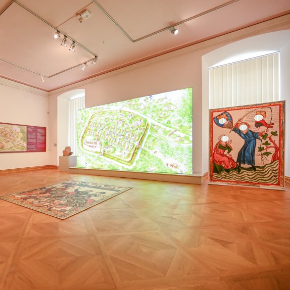 Ansicht eines Ausstellungsraumes mit großem Bodenpuzzle (Motiv aus "Codex Manesse"), Selfiewand (Motiv aus "Codex Manesse") und einer großen Leuchtwand, die eine Ansicht Ettlingens im Mittelalter zeigt.