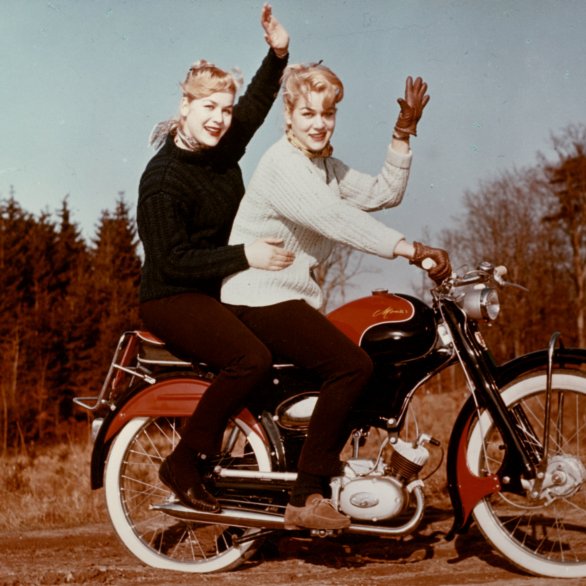 Farbfoto von winkenden weiblichen Zwillingen auf einem scwharz-roten Moped.