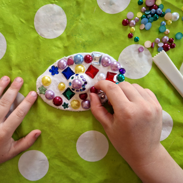 Kinderhände verzieren eine Tonscheibe mit bunten Glasperlen