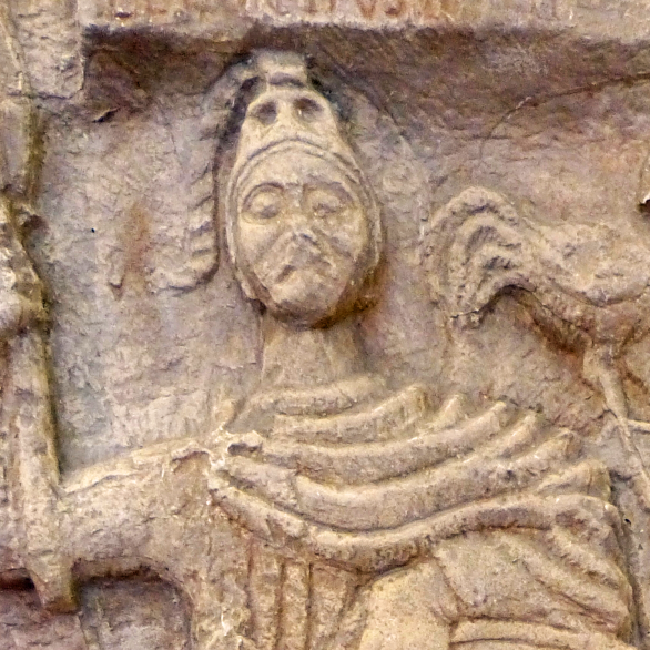 Person mit Umhang und Kopfbedeckung und seitlich einem Hahn