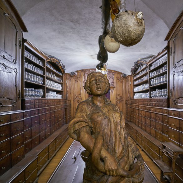 Holzgeschnitzte Figur auf einem Apothekentisch, links und rechts das Mobiliar der Innsbrucker Hofapotheke. An der Decke hängt ein ausgestopfter Kugelfisch
