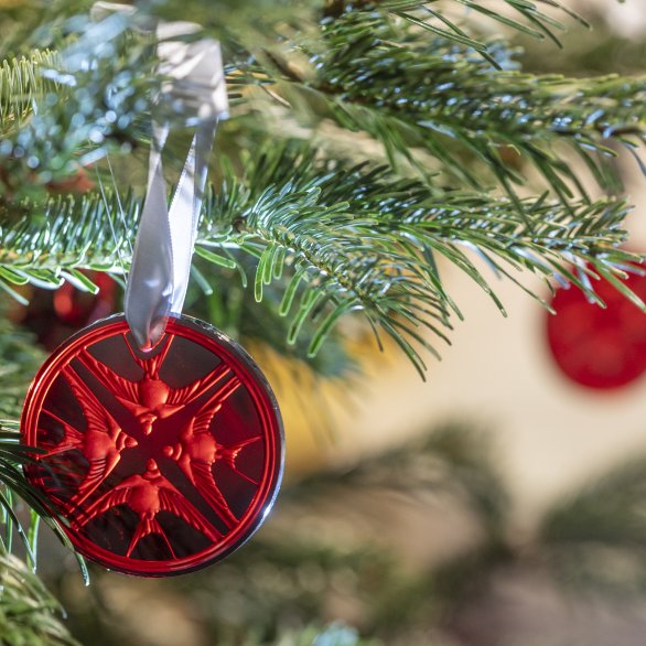 Ornement de Noël Lalique rond en cristal rouge représentant 4 hirondelles accroché à une branche de sapin avec un ruban clair