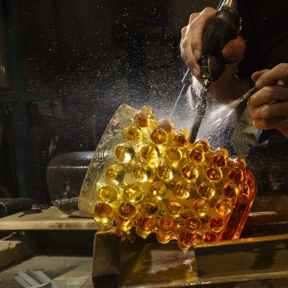 un vase en cristal ambre avec des motifs de boules est en train d'être taillé par une main qui tient un outil et une autre qui tient un jet d'eau