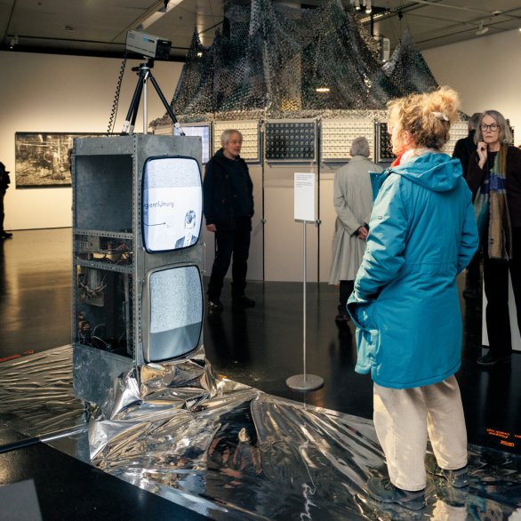 Besucher:innen der Ausstellung stehen vor zwei übereinander gestapelten Fernsehern, auf denen ein Video läuft.