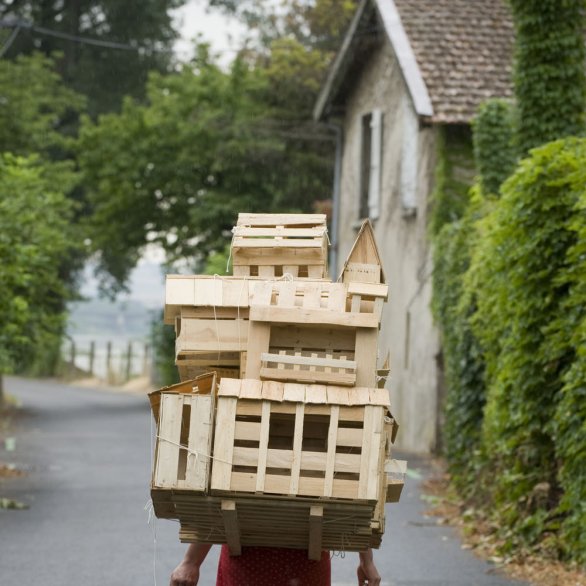 Werk der Künstler Gabriela Oberkofler, Fotografie, auf der eine als Frau gelesene Person zu sehen ist, die ein Modell eines Hauses auf dem Rücken trägt