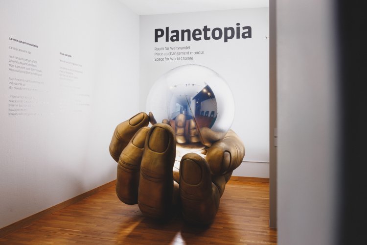 Eine grosse Hand hält eine silberne Kugel, worin wir Betrachtenden uns spiegelt. Es ist der Eingang zur Ausstellung "Planetopia - Raum für Weltwandel"