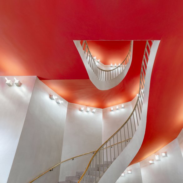 Blick nach oben in ein geschwungenes Treppenhaus: Weiße Wände und Geländer rahmen leuchtend rot-orange Deckenflächen.