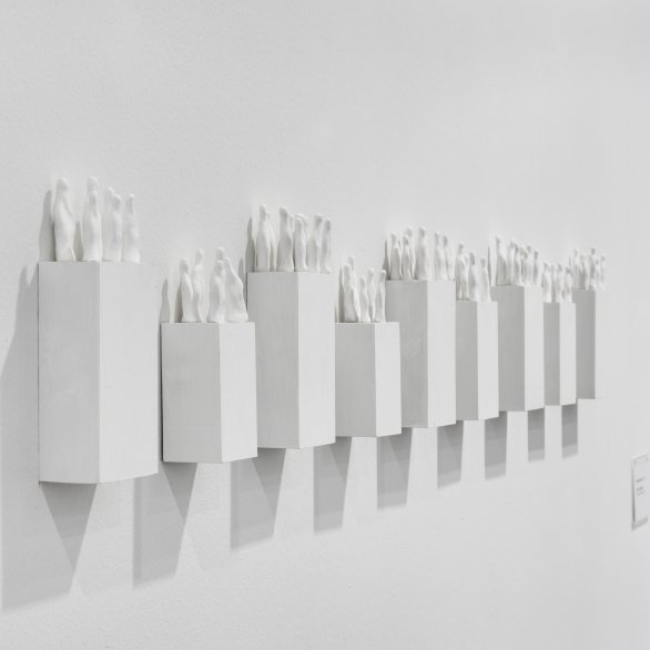 Weiße Skulpturarbeit mit abstrahierten Figuren auf rechteckigen Sockeln in einer Wandreihe.