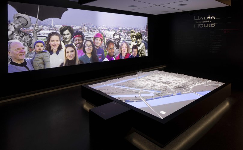 Das interaktive Stadtmodell zeigt eine Projektion auf den nachgebauten Stadtplan von Mannheim. Im Hintergrund ist eine Menschenmenge projeziert, die vor Mannheims Stadtkulisse steht.