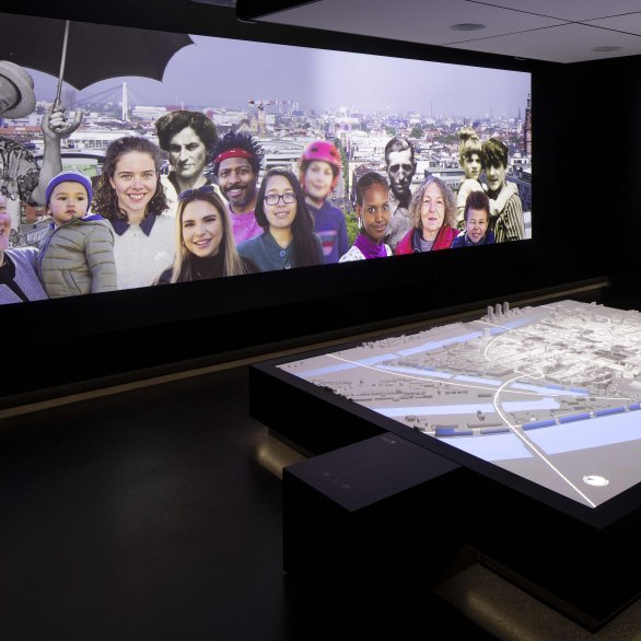 Das interaktive Stadtmodell zeigt eine Projektion auf den nachgebauten Stadtplan von Mannheim. Im Hintergrund ist eine Menschenmenge projeziert, die vor Mannheims Stadtkulisse steht.