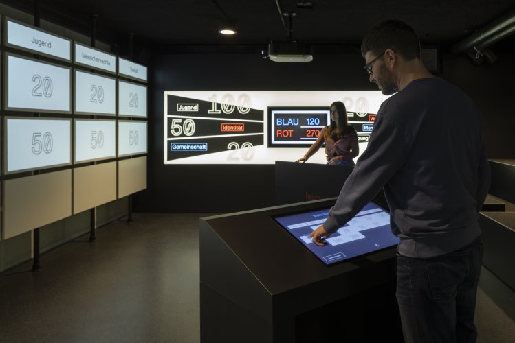 Blick in den Raum, in dem sich das interaktive, digitale Spiel "Democrady" befindet. Zwei Personen stehen seitlich vor einer großen Leinwand  und blicken auf einen kleinen Monitor.
