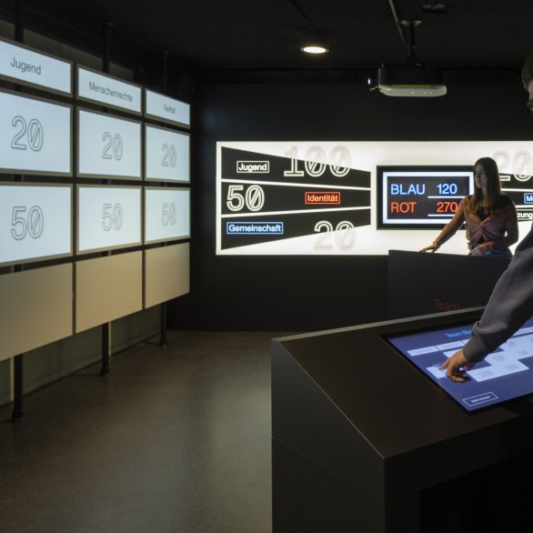 Blick in den Bunkerraum. Große Displays zeigen Kinder, die in Trümmern spielen.